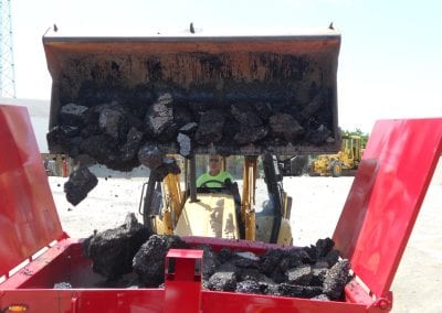 A backhoe operator using the front bucket to load recycled asphalt chunks into the hot box.